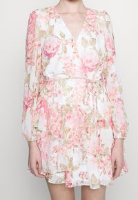 Kukkapuku vaaleassa, jossa on vaaleanpunaisia ruusuja, syvä V-mallinen kaula-aukko, pitkä puffihihna ja nyöritetty vyötärö. Aaltoileva kerroksellinen helma.