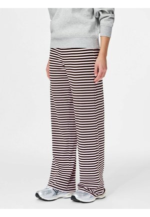 Personne portant un pantalon large et ample à rayures horizontales noires et roses, avec des chaussures de sport argentées, debout sur un fond blanc.