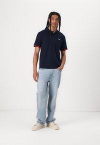 Navy polo shirt with red accents, short sleeves, and a small logo; paired with light blue baggy jeans and white sneakers.