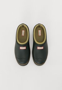 Scarpe slip-on nere in gomma con punta rotonda, fodera verde oliva e un piccolo logo rosso e bianco sul lato. Suola marrone e testurizzata.