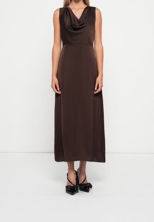 Femme portant une robe marron foncé sans manches, longueur cheville, avec un col drapé et des escarpins noirs à bout pointu avec des nœuds.