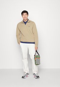 Fleece bej cu fermoar la jumătate, cămașă albastră dedesubt, pantaloni cargo albi și adidași gri/negru. Ținând o geantă verde cu accente portocalii.