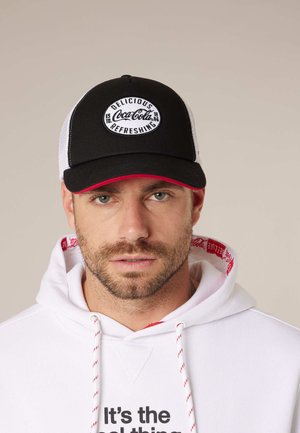 Homme portant une casquette Coca-Cola noire et blanche et un sweat à capuche blanc avec texte rouge et noir, regardant devant lui sur un fond uni.