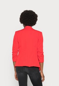 Blazer rouge avec une coupe ajustée et un dos légèrement évasé. Il se caractérise par des manches longues froncées et un col cranté. Le tissu semble lisse et structuré.