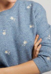 Pull en tricot bleu clair avec un col rond, orné de motifs floraux brodés blancs et de poignets côtelés pour une touche de texture.