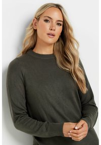 Maglione verde scuro realizzato in un morbido cotone testurizzato; presenta un collo rotondo e maniche lunghe con polsini a coste. Modella mostrata su uno sfondo neutro.