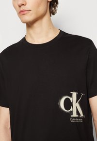 Černé bavlněné tričko s kulatým výstřihem a krátkými rukávy, s bílým sprejovaným logem "CK" a názvem značky na hrudi.