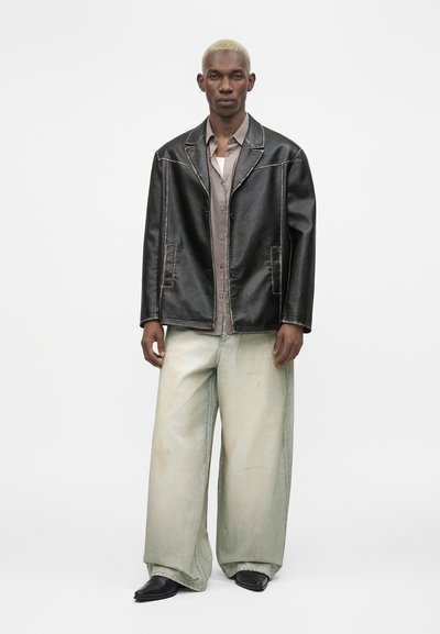 Άνδρας μοντέλο με κοντά ξανθά μαλλιά, φοράει υπερμεγέθη μαύρο δερμάτινο σακάκι, γκρι πουκάμισο με κουμπιά, φαρδιά ξεβαμμένα ανοιχτόχρωμα τζιν και μαύρα μυτερά παπούτσια.