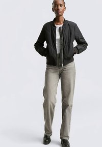 Črna prešita bomber jakna z zadrgo, rebrastimi manšetami in belo majico spodaj; kombinirana s svetlo sivimi jeans hlačami z ravnimi hlačnicami.