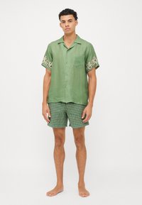 Camisa verde de manga corta con botones y bordado en espiral en las mangas, combinada con pantalones cortos de cuadros verdes. La modelo está descalza.