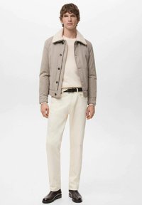 Beige jacka med fårskinnskrage, knäppt fram, creamfärgad tröja, ljusa byxor och mörka läderstövlar. Enkel, modern design.