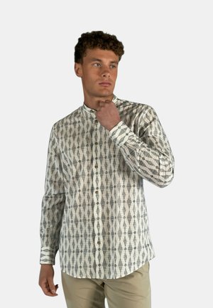 Giovane uomo che sistema il colletto, indossa una camicia a maniche lunghe con fantasia e pantaloni beige, guarda leggermente di lato.