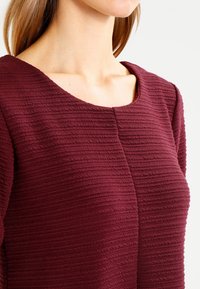 Burgundy texturerad topp med rundad halsringning, som har vertikal ribbning och en figursydd siluett, vilket framhäver dess unika tygdetalj.