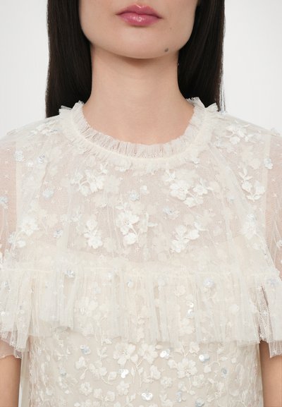 Läpinäkyvä kermaväri blouse, jossa on kukka-embellishments, paljetit ja rypytetty kaula-aukko. Kevyt rakenne korostaa herkkiä muotoiluelementtejä.