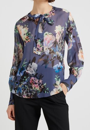 Frau trägt eine durchsichtige marineblaue Bluse mit mehrfarbigem Blumenmuster und einer gebundenen Schleife am Ausschnitt, kombiniert mit schwarzen Hosen.
