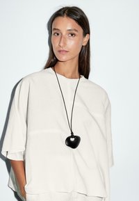 Une jeune femme aux longs cheveux raides porte un haut blanc ample et un collier avec un pendentif noir distinctif sur un fond pâle.