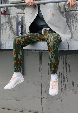 Joggers mimetici in verde e marrone, realizzati in tessuto morbido, abbinati a sneaker bianche con dettagli rosa e suole testurizzate. Stile urbano casual.