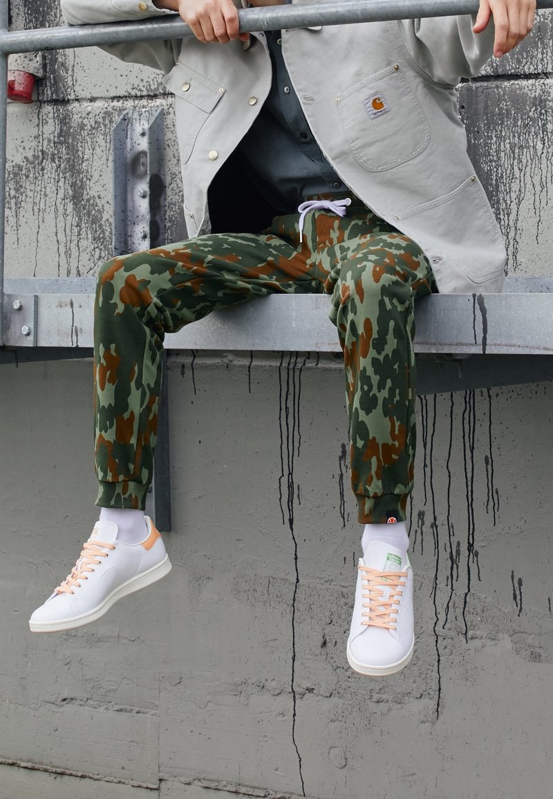 Camo joggers in green and brown, made of soft fabric, paired with white sneakers featuring pink accents and textured soles. Casual urban style.