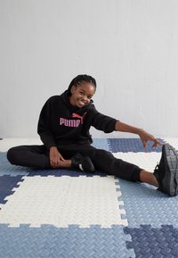 Sudadera negra con el logo rosa de Puma, pantalones negros a juego y zapatillas deportivas negras. Sentado sobre azulejos de espuma entrelazados en azul y blanco.