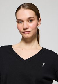 Svart v-ringad tröja i lätt stickad kvalitet, med en liten, broderad logotyp på vänster bröst. Slät textur och avslappnad design.