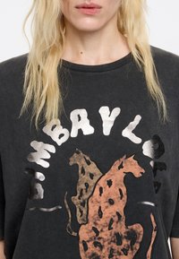 Pelēks pārāk liels T-krekls ar divu stilizētu leopardu grafiku zemes toņos, ar vārdu "BAYLOR" drukātu drosmīgās sudrabainās burtiem.