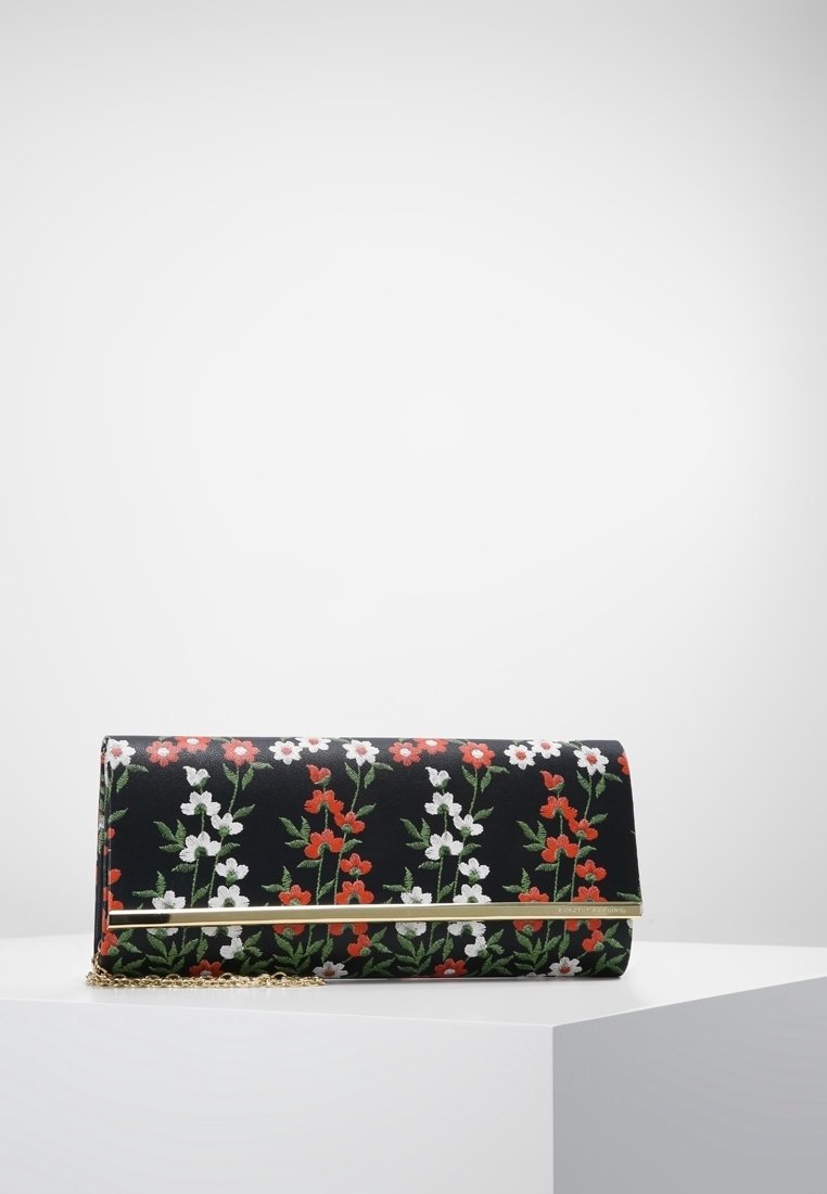 Pochette noire avec broderies florales rouges et blanches, bordure dorée le long du rabat, et une délicate chaîne dorée en bandoulière, posée sur un piédestal blanc.