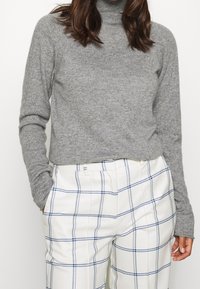 Grauer Rollkragenpullover mit langen Ärmeln, kombiniert mit weißen Karo-Hosen mit blauen Linien, die einen strukturierten Stoff betonen.