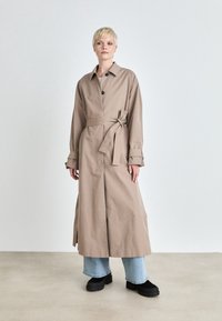 Osoba stojící v béžovém trenčkotě s páskem, světle modrých džínách a černých robustních botách na pozadí světlého jednobarevného pozadí.