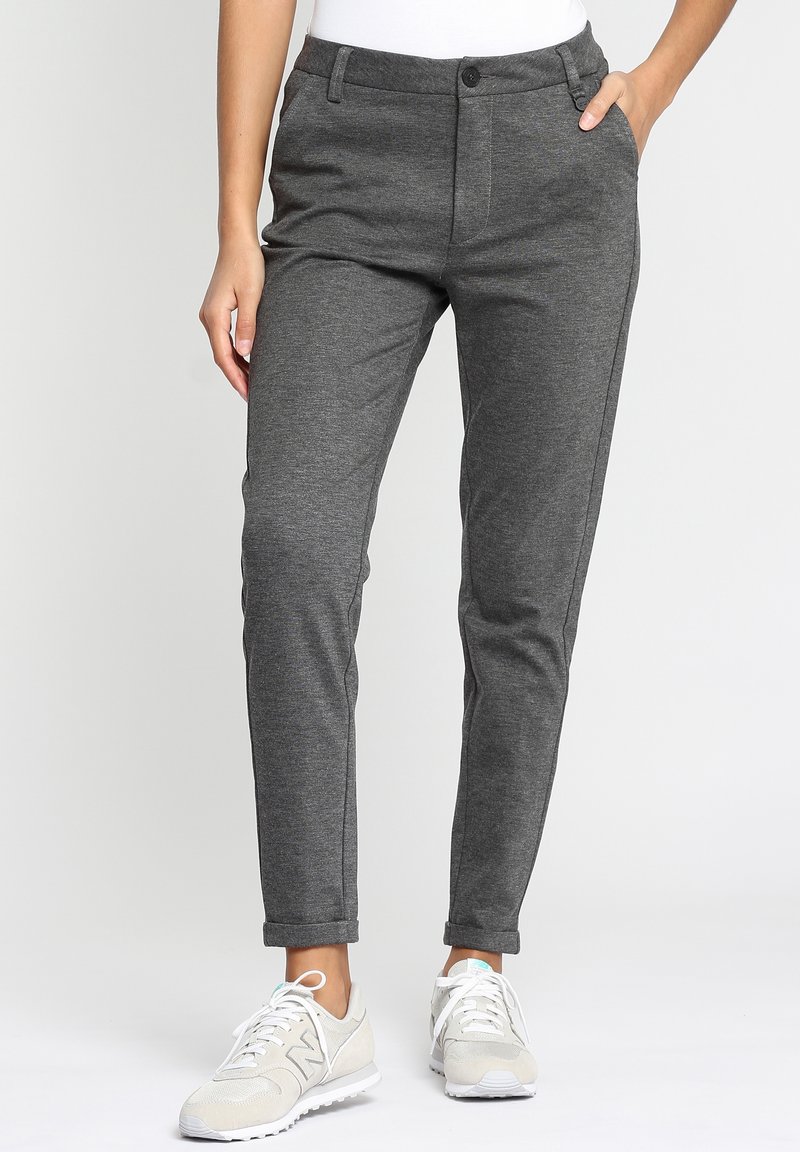 Femme portant un pantalon gris slim avec les ourlets retroussés et des baskets blanches, debout avec une main dans la poche sur un fond uni.