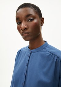 Blaue Bluse mit Knopfverschluss, aus weichem Stoff, mit leichter Raffung an den Schultern und glatter Textur.