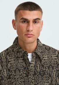 Črna srajca s pritiski in zapletenimi zlatimi paisley vzorci, s ovratnikom in žepom na prsih, nosi se čez belo srajco.