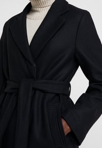 Cappotto nero con vita cintrata, colletto a scialle e due tasche laterali; realizzato in tessuto testurizzato con una finitura morbida.