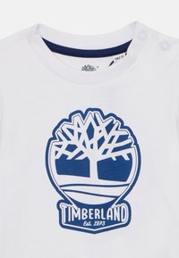 Valkoinen puuvillainen t-paita, jossa sininen Timberland-logo, jossa on puu ja maisemakuva. Olkavarressa on kaksi painonappia.