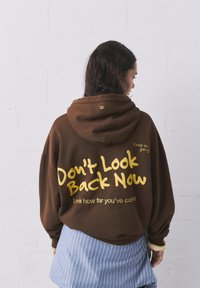 Hnedý veľký mikinový sveter s nápisom zlato: "Neobzeraj sa späť" a "Pozri, ako ďaleko si sa dostal." Obsahuje kapucňu a rebrované manžety.