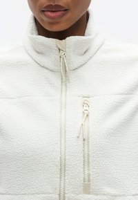 Λευκό fleece μπουφάν με ψηλό γιακά, που διαθέτει τσέπη με φερμουάρ και υφή φινιρίσματος. Τα φερμουάρ είναι σε ανοιχτό μπεζ χρώμα.