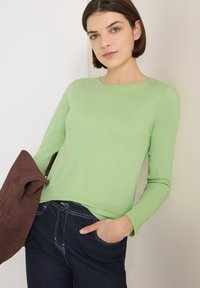 Top de manga larga color verde claro hecho de tela suave, combinado con vaqueros oscuros. Presenta un escote redondo y un ajuste relajado, con una chaqueta marrón.