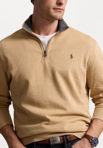 Beige pullover tröja med halv dragkedja, med en grå inre krage och en liten broderad logotyp på bröstet. Mjuk textur.
