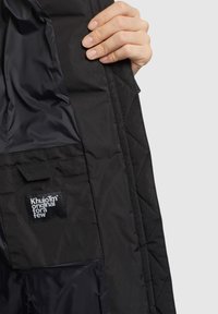 Sort quiltet jakke, der viser det indvendige foer med en blank finish og et mærke af stof med teksten "Khujo original for a few."