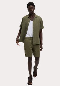 Camisa de botones de manga corta con un patrón verde, camiseta blanca sin mangas, pantalones cortos de carga color oliva y sandalias marrones, que muestran un ajuste relajado y un estilo casual.