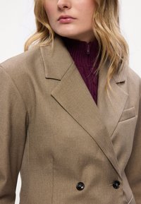 Blazer castanho de duas fileiras de botões com lapelas recortadas. Apresenta um tecido texturizado e um bolso no peito, acompanhado de um pulôver com fecho de correr bordeaux por baixo.