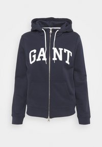 Felpa con zip navy in tessuto misto cotone. Presenta una grande stampa bianca "GANT" sul davanti, cappuccio con cordino e tasche frontali.