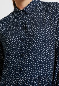 Abito blu navy con bottoni, caratterizzato da un piccolo motivo floreale bianco, tessuto testurizzato e un orlo a balze. 