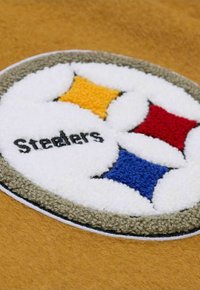 Runder, bestickter Aufnäher mit weißem Hintergrund, schwarzem Text "Steelers" und bunten Rautendesigns in Gelb, Rot und Blau auf einem hellbraunen Stoff.