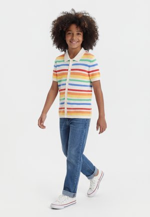 Dieťa kučeravých vlasov, ktoré chodí, oblečené v krátkom svetríku s golierom a farebnými vodorovnými pruhmi, modré rifle a biele tenisky.