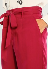 Pantalon fuchsia taille haute avec ceinture nouée, poches latérales et texture lisse. La taille est froncée pour un ajustement plus cintré.
