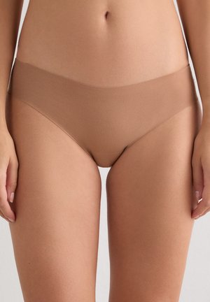 Mujer con ropa interior tipo hipster sin costuras y color nude contra un fondo neutro, mostrando torso y parte superior de los muslos.
