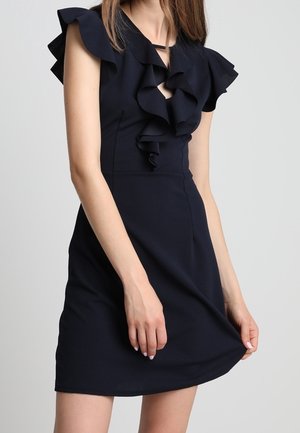 Kvinde iført sort ærmeløs kjole med flæset halsudskæring og kant, der holder skørtet let ud mod en ens grå baggrund.