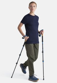 Námornické tričko s krátkým rukávem, olivově zelené kalhoty a modré turistické boty. Uživatel drží černé trekingové hole s modrými akcenty a korkovými rukojeťmi.