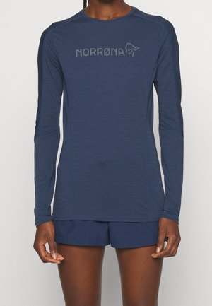 Osoba nosząca dopasowaną, granatową bluzę z długim rękawem z logo „NORRØNA” oraz pasujące granatowe szorty, stojąca na jednolitym tle.