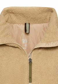 Beige fleecejakke med høj krave, halv-zip front, quiltet inderside og en tyk tekstur. Har en løkke til ophængning og en grøn zip-puller.